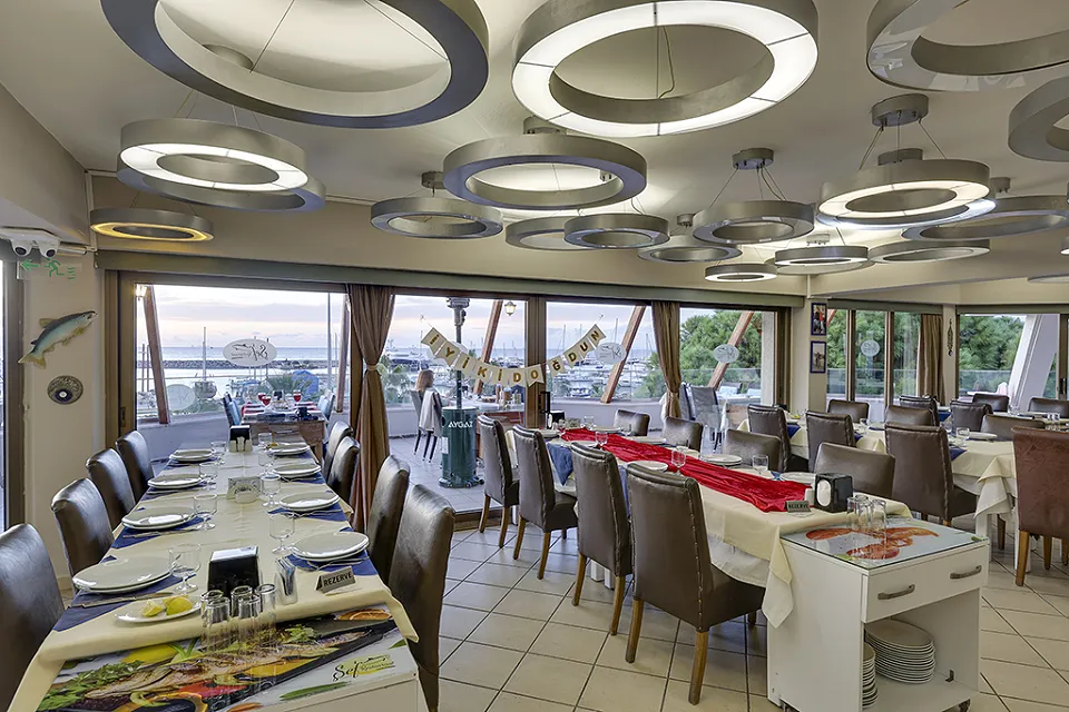 Long tables with red runners and wide windows facing the sea in the indoor dining room with ring chandeliers