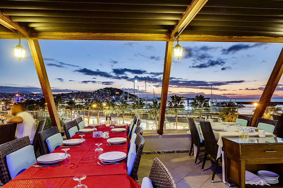 Tables with red tablecloths under a wooden pergola hung with lanterns, with city lights at dusk
