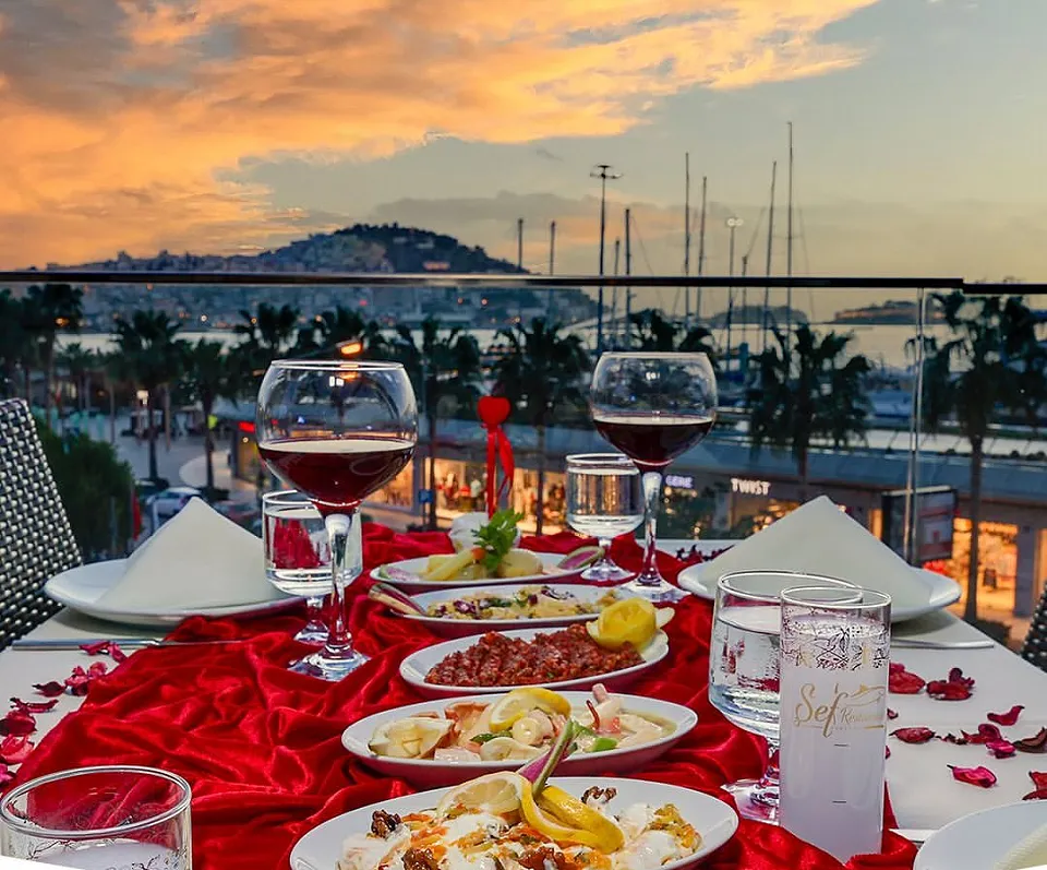 A red tablecloth scattered with rose petals, wine glasses and meze on the terrace overlooking the marina at sunset