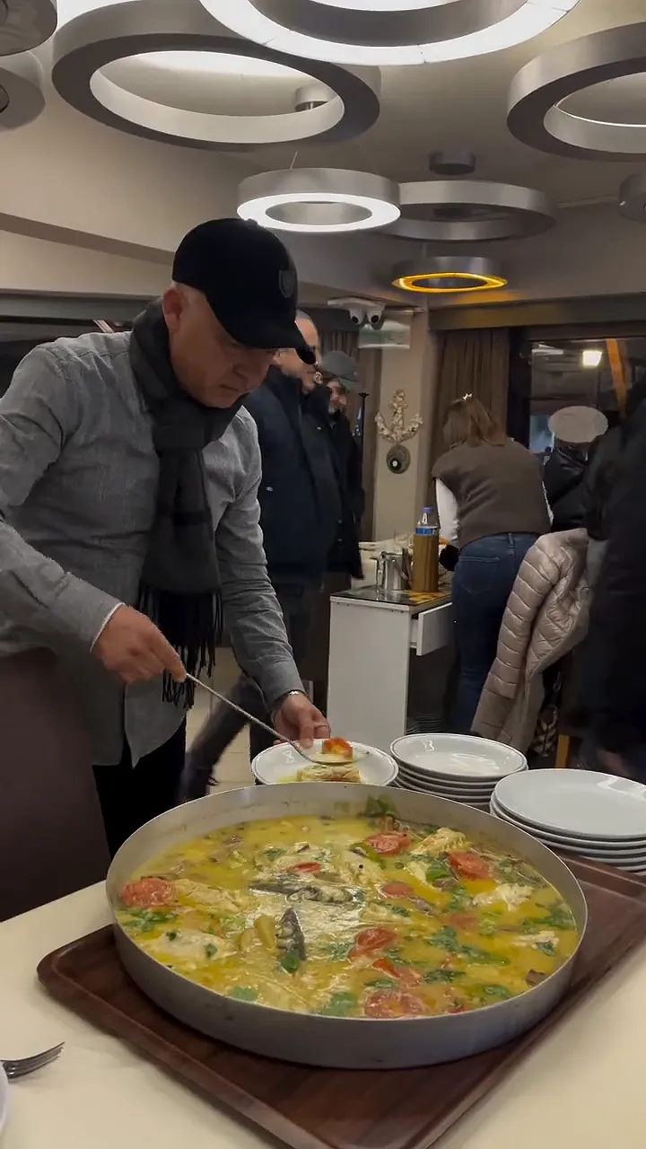 Video: fish poached with tomatoes being served from a large tray onto plates in a dining room with ring chandeliers